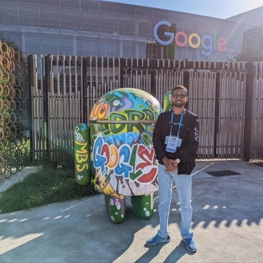 A person standing next to a colorful graffiti-painted Android statue, with the Google headquarters in the background. The person is wearing a lanyard on a sunny day.